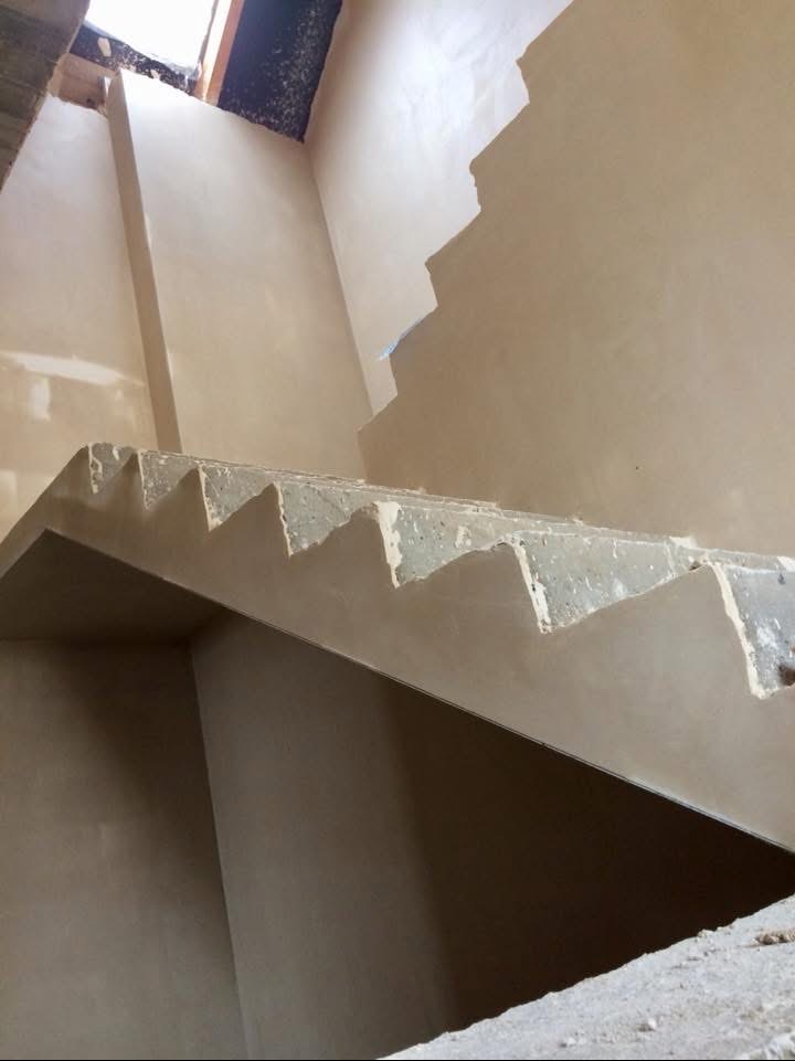 Wnętrze klatki schodowej w trakcie prac wykończeniowych. Ściany i schody pokryte świeżym, jasnym tynkiem. Widoczne niedoskonałości i surowość materiału.