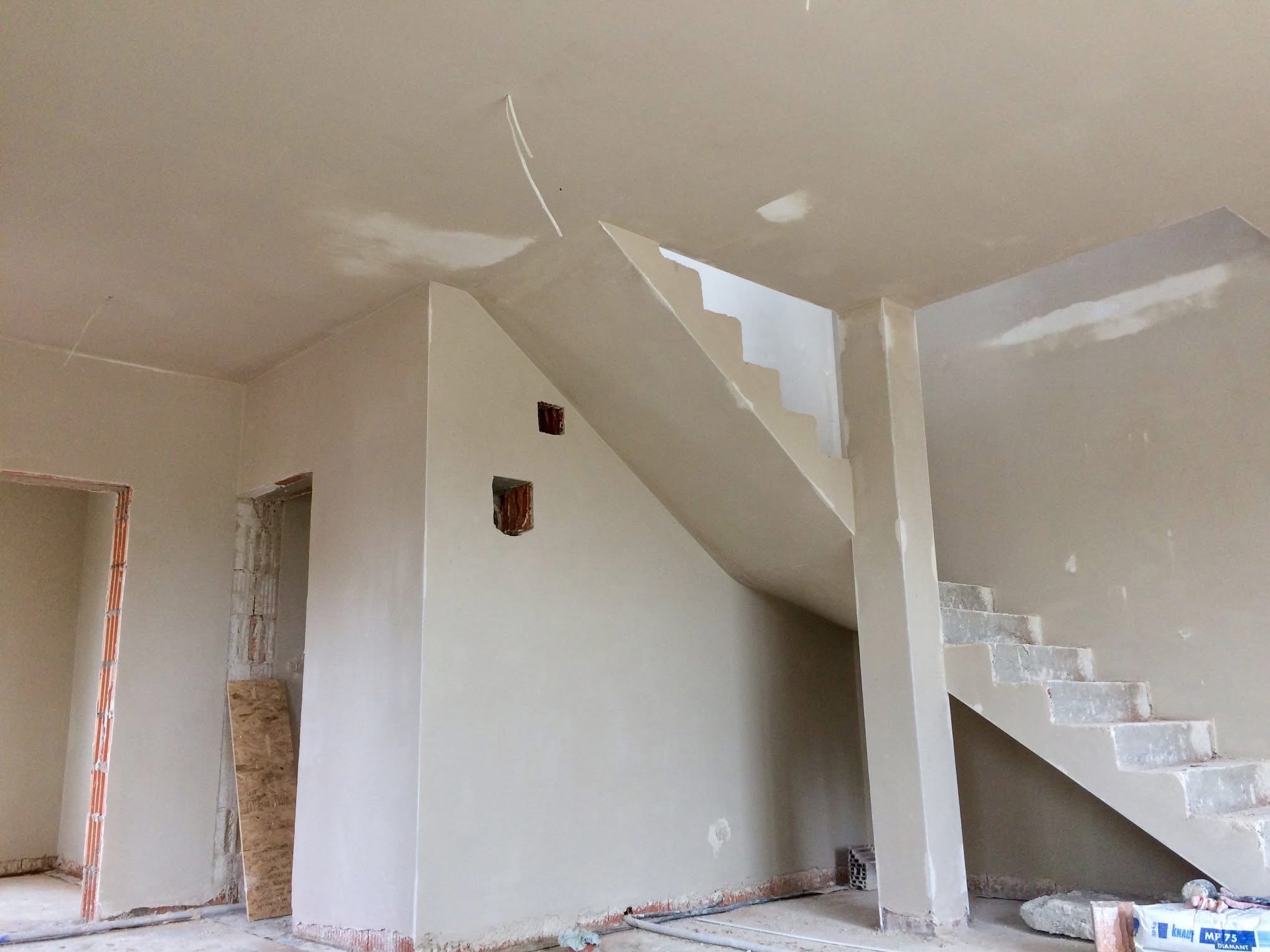 Wnętrze w trakcie tynkowania maszynowego: gładkie, jasne ściany, schody w surowym stanie, widoczne puszki elektryczne i przewody. Na podłodze resztki materiałów budowlanych.