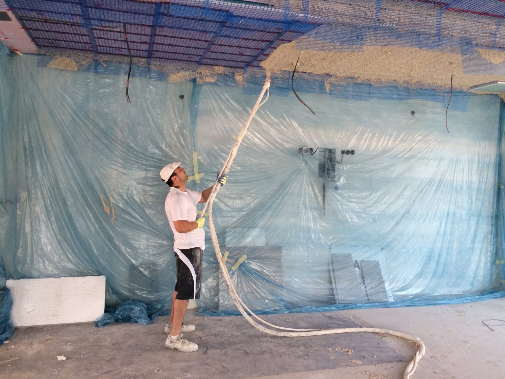 Pracownik w trakcie natrysku tynku na sufit, zabezpieczony folią. Widoczne przewody i siatka na suficie. Ujęcie z dołu, prezentujące etap prac wykończeniowych.