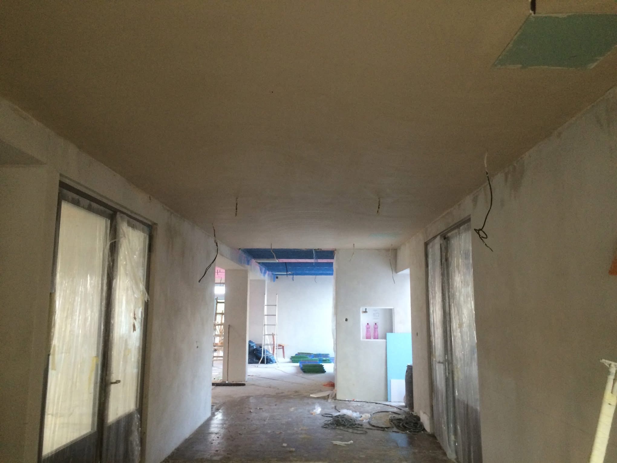 Wnętrze w trakcie prac wykończeniowych: świeżo położone tynki na ścianach i suficie, zabezpieczone folią drzwi, widoczne przewody i elementy instalacji.