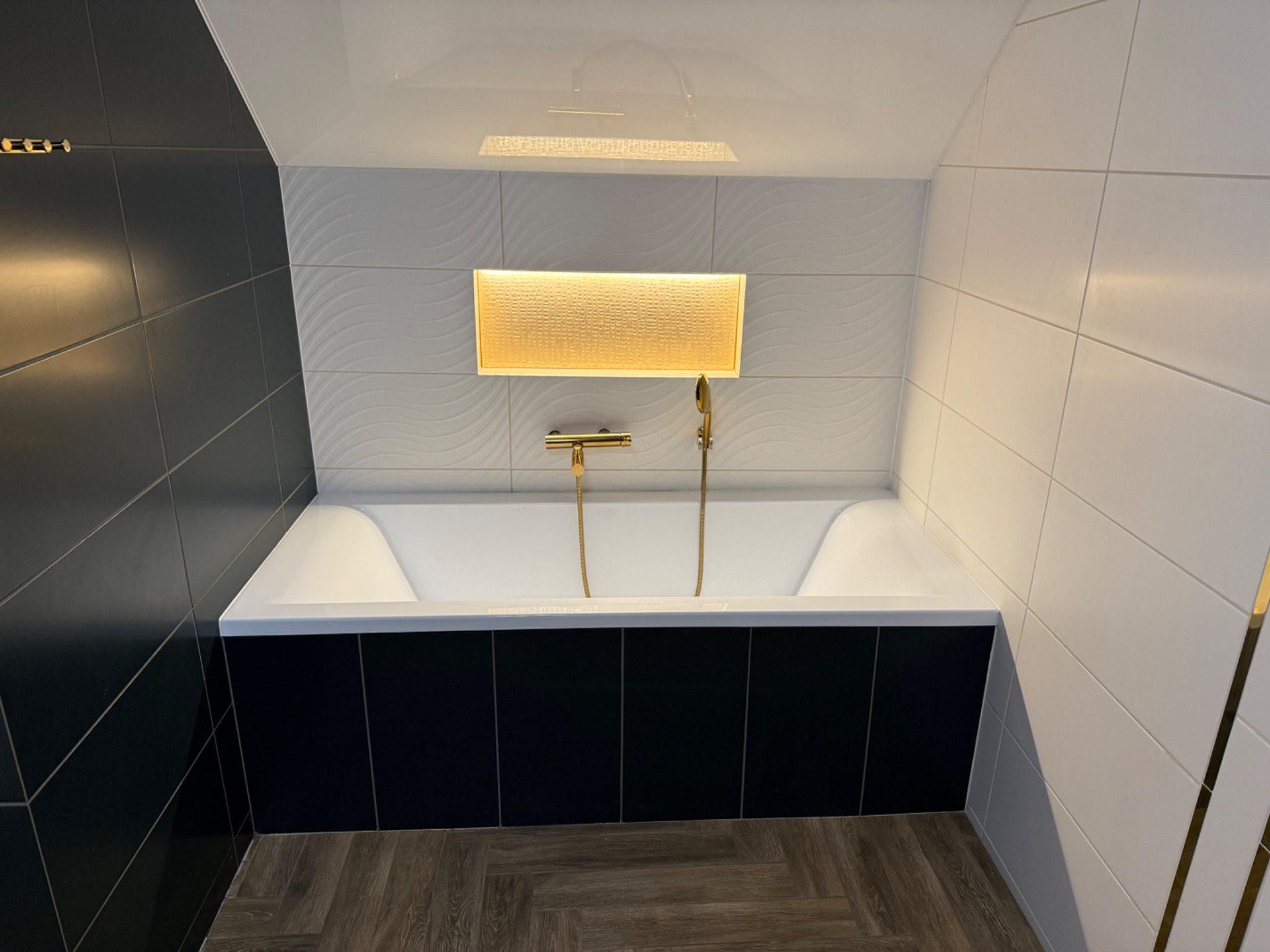 Nowoczesna łazienka z wanną obudowaną czarnymi płytkami, złota armatura i oświetlenie, białe płytki na ścianach z delikatnym wzorem fal. Parkiet na podłodze.