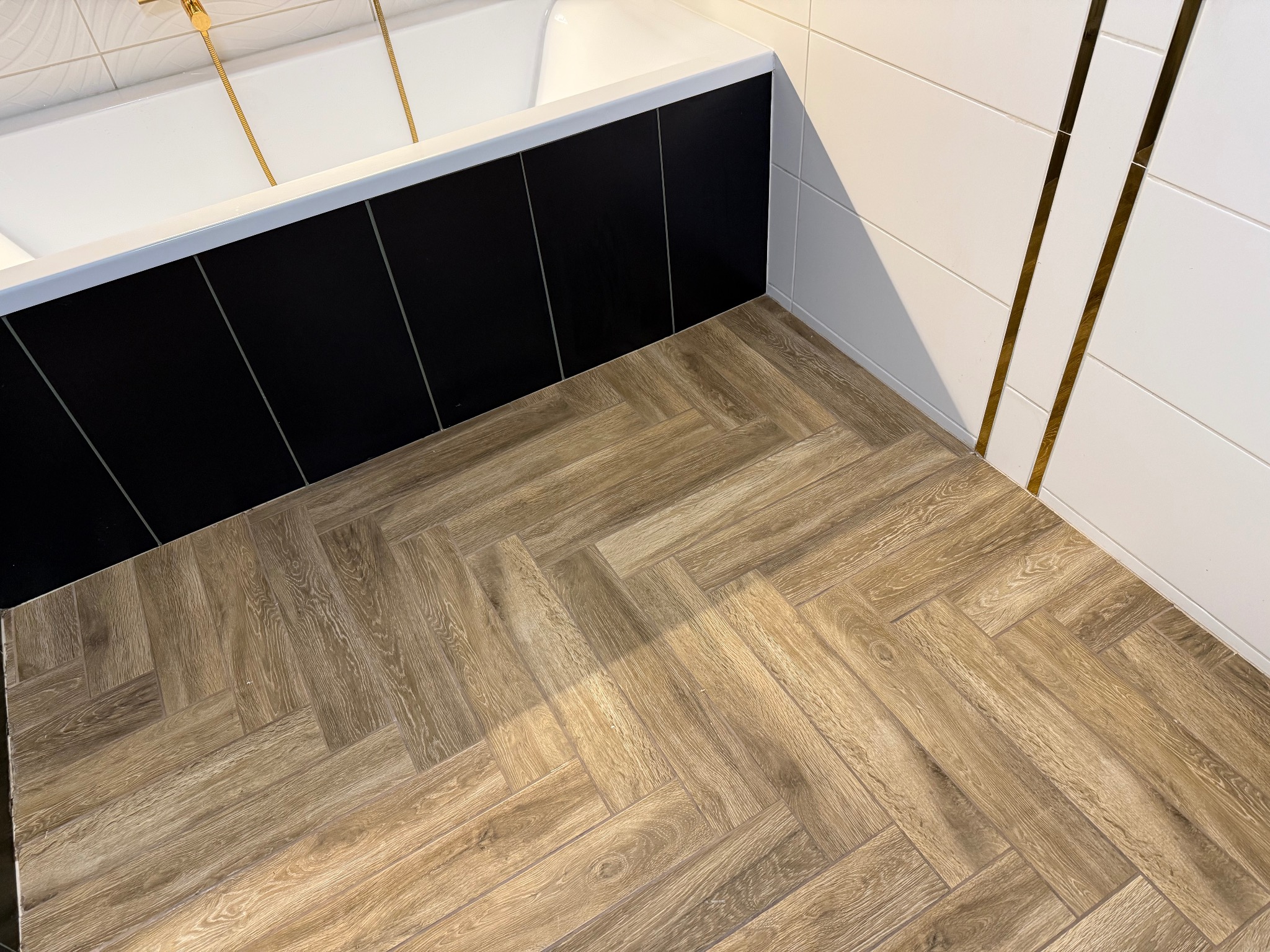 Ukończona łazienka z parkietem ułożonym w jodełkę, czarną obudową wanny i białymi płytkami z złotymi paskami. Nowoczesny design.