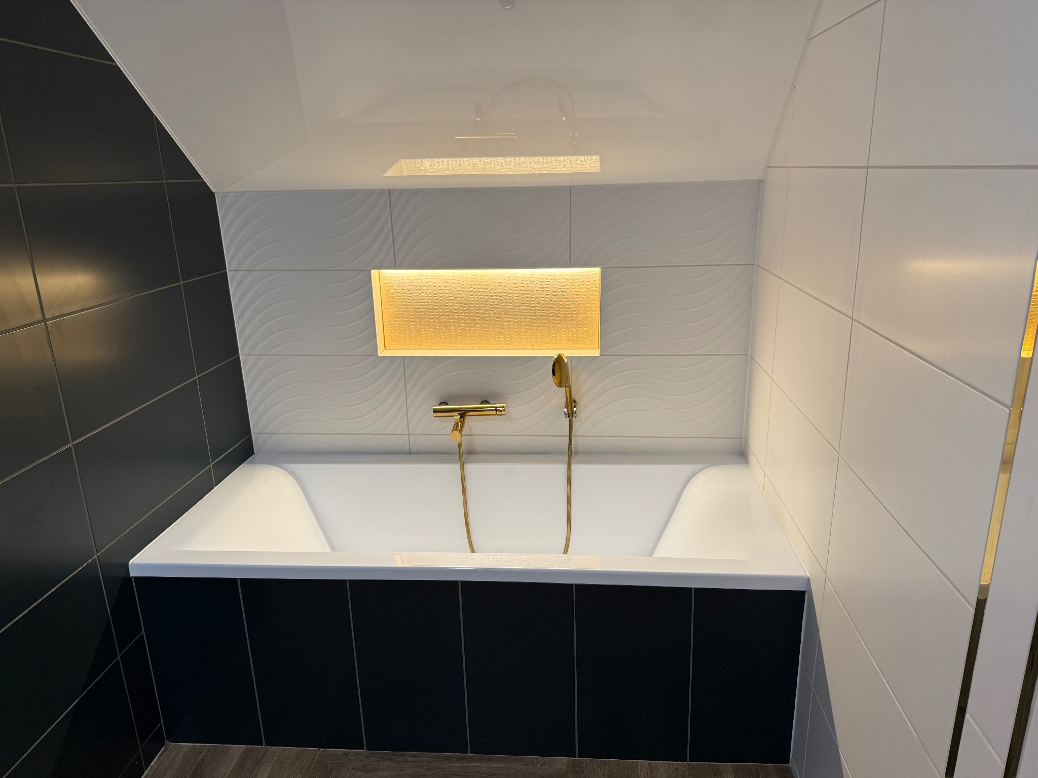 Nowoczesna łazienka z białą wanną obudowaną czarnymi płytkami, oświetlona listwą LED. Białe płytki z delikatnym wzorem na ścianie i błyszczący sufit.