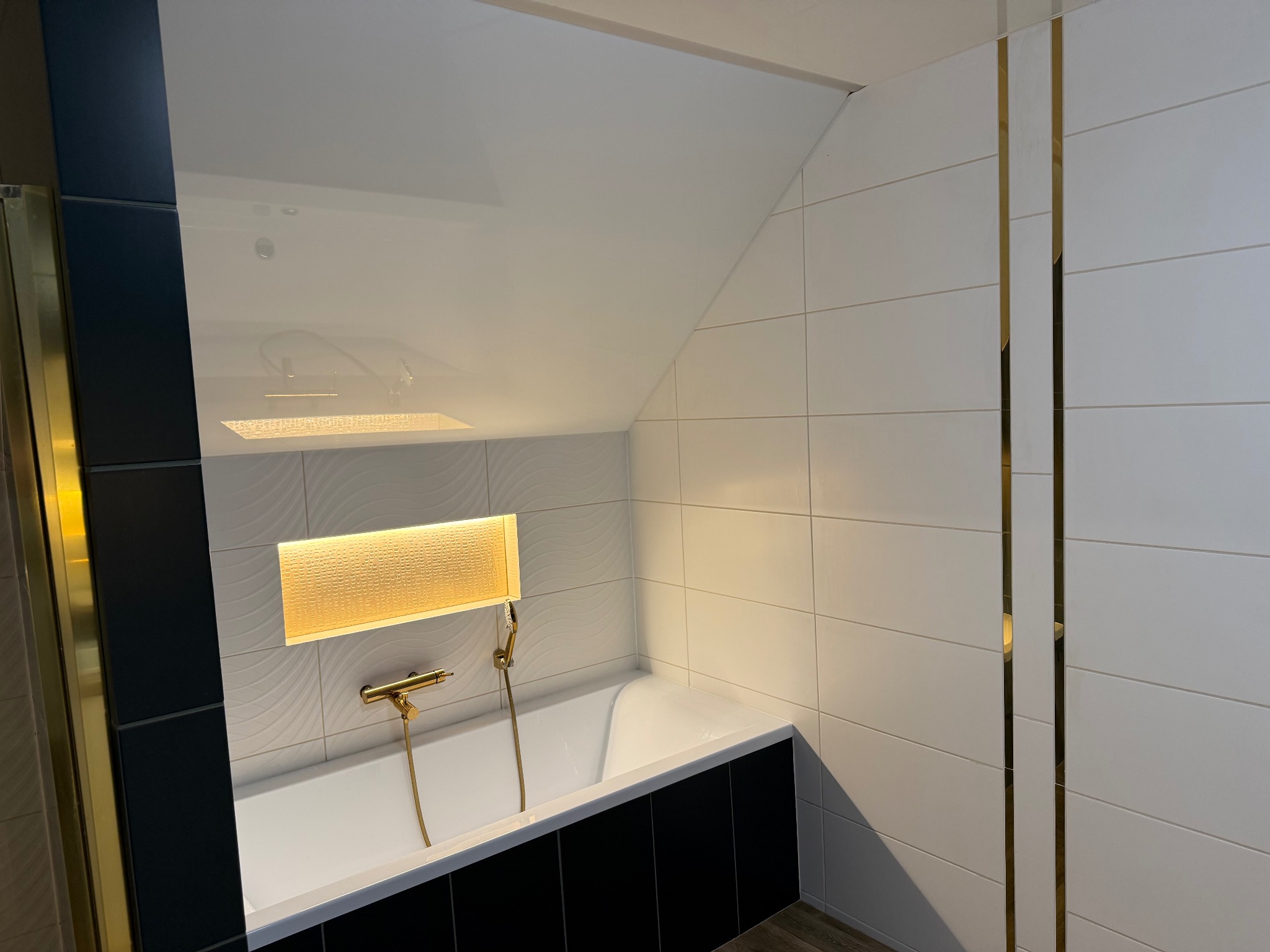 Nowoczesna łazienka z biało-czarnymi płytkami, złotymi akcentami i podświetlaną wnęką nad wanną. Minimalistyczny design z eleganckimi detalami.