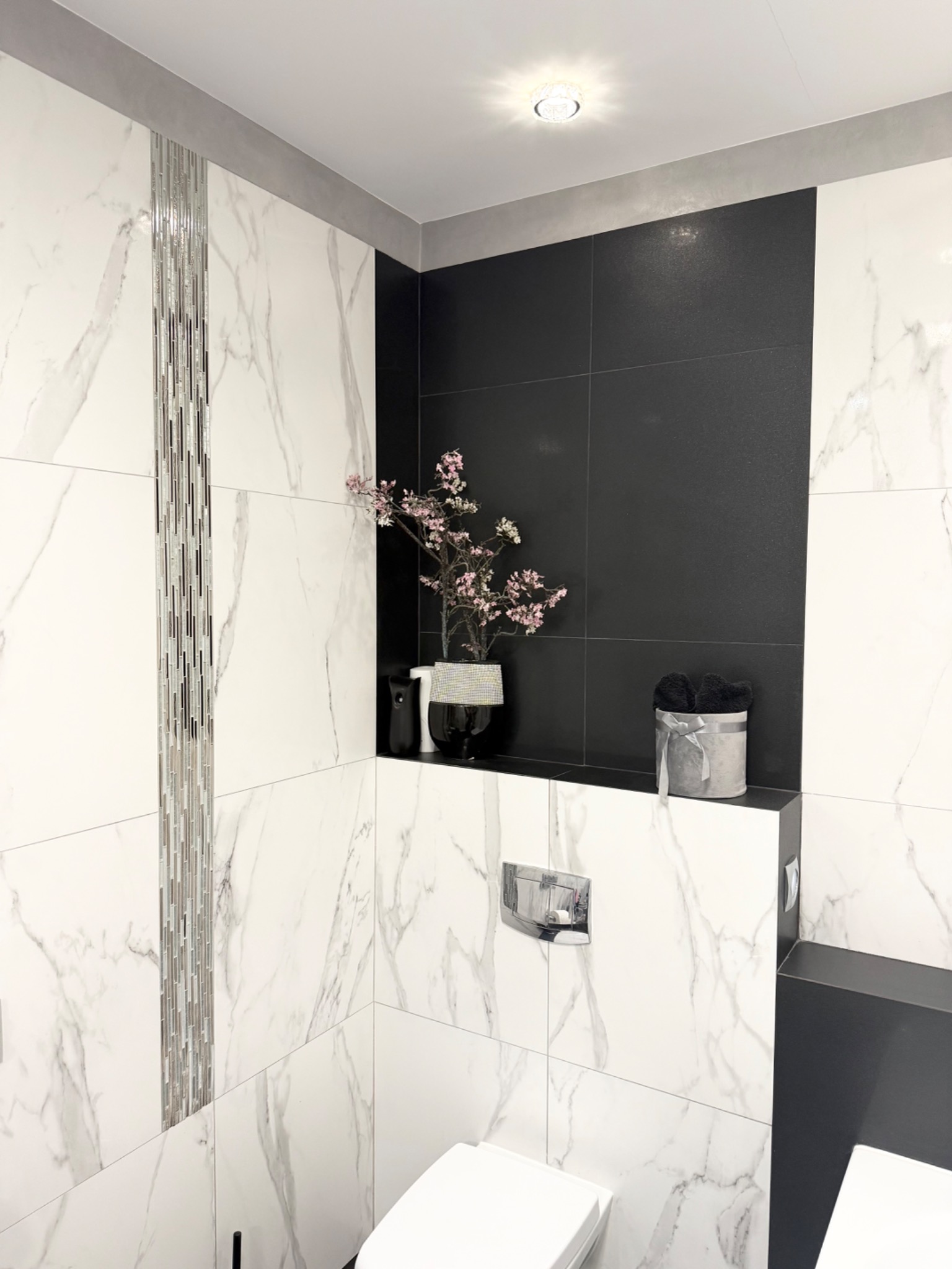 Elegancka łazienka z białymi płytkami imitującymi marmur, czarnymi akcentami i dekoracyjnymi elementami, takimi jak kwiaty i ozdobne pudełko.