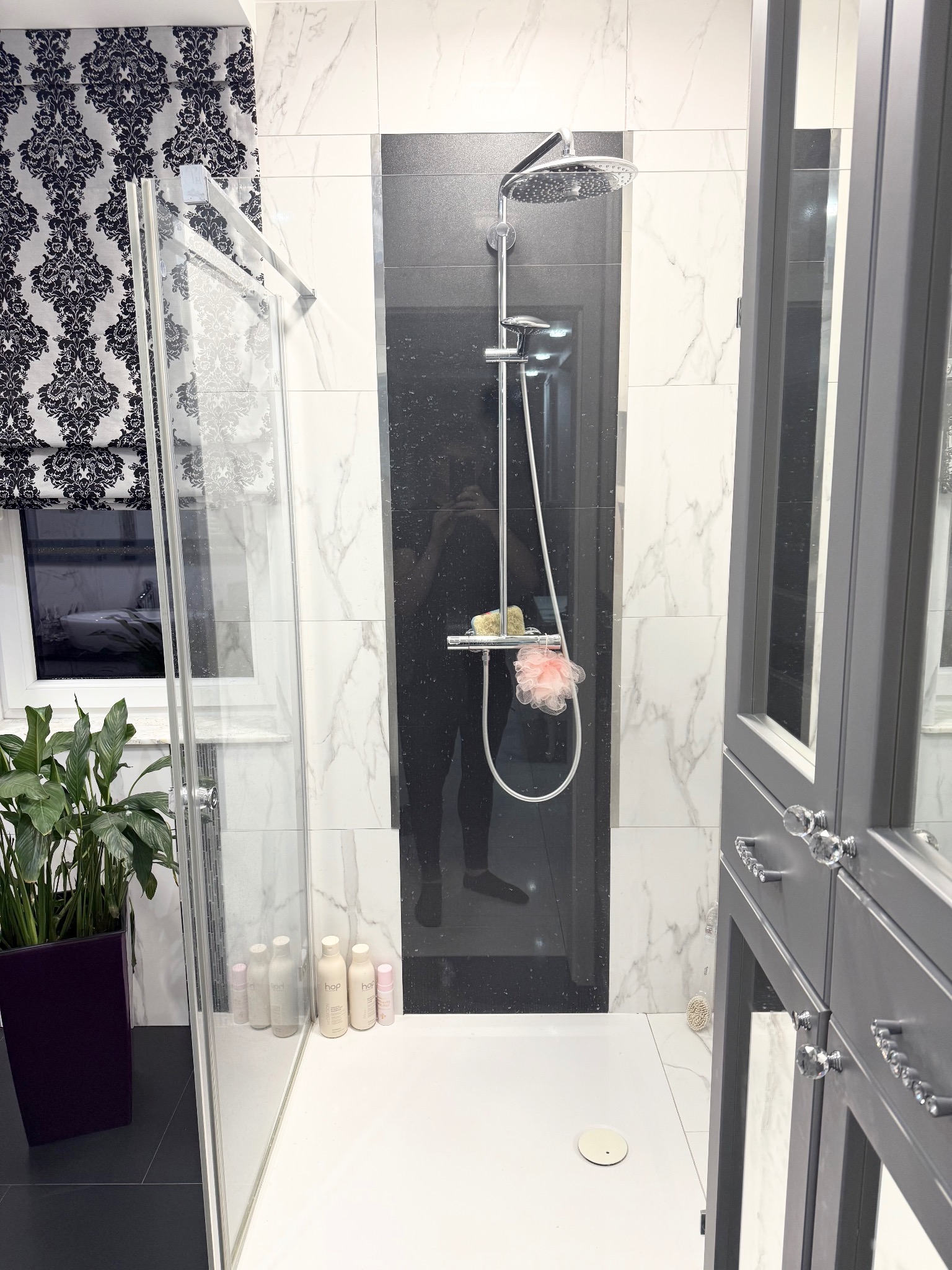 Nowoczesna kabina prysznicowa z deszczownicą, czarno-białe płytki imitujące marmur, szara szafka łazienkowa z kryształowymi uchwytami i roślina w doniczce.