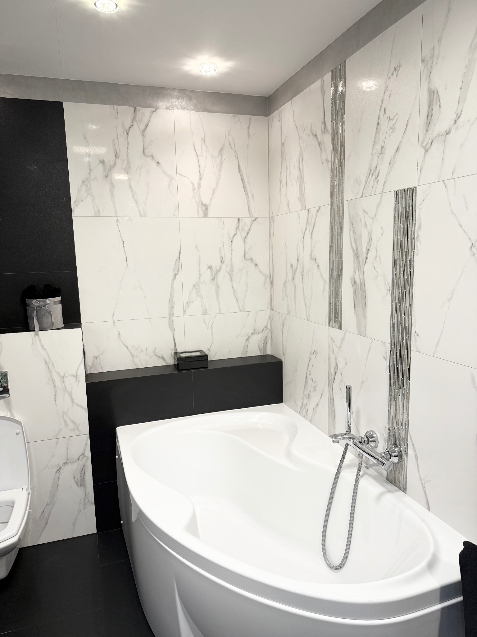 Nowoczesna łazienka z białą wanną, marmurowymi płytkami i czarnymi akcentami. Bateria wannowa z chromu. Oświetlenie punktowe. Minimalistyczny design.