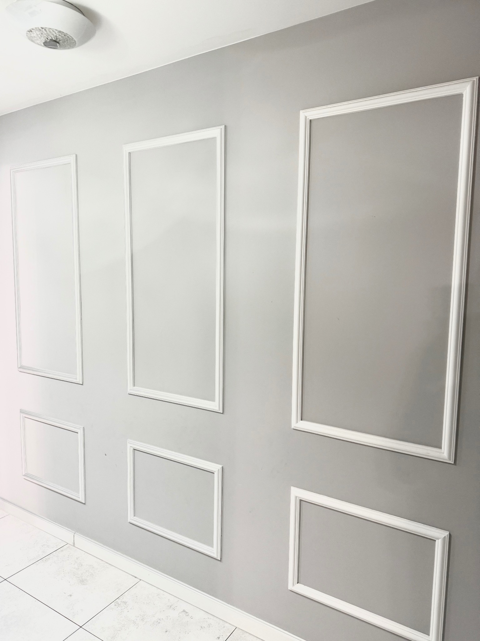 Szara ściana z dekoracyjnymi, białymi ramkami w różnych rozmiarach i kształtach. Widoczny fragment podłogi z białych płytek i lampa sufitowa.