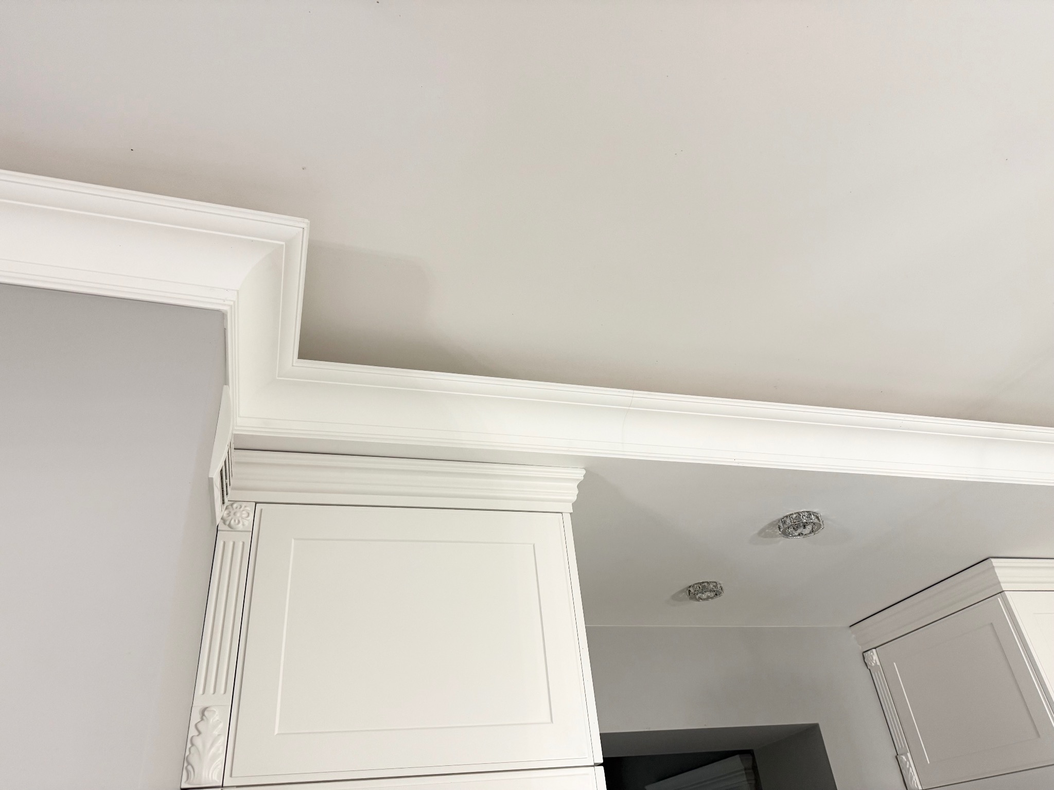 Białe, klasyczne szafki kuchenne pod sufitem z ozdobnymi listwami. Widoczne oświetlenie sufitowe i szara ściana. Ujęcie z dołu, podkreślające detale wykończenia.