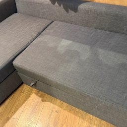 Górska Opieka Nieruchomości - Szara, rozłożona kanapa narożna z tkaniny na drewnianej podłodze. Widoczny mechanizm rozkładania i faktura materiału. Ujęcie z góry, naturalne światło.