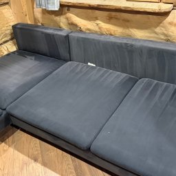 Górska Opieka Nieruchomości - Szara, narożna kanapa po czyszczeniu, z widocznymi śladami po odkurzaczu piorącym, w drewnianym wnętrzu. Widoczne jasne smugi na ciemnej tapicerce.