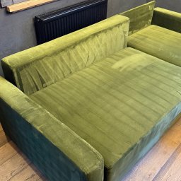 Górska Opieka Nieruchomości - Zielona, aksamitna sofa modułowa na drewnianej podłodze, z widocznym grzejnikiem i dekoracyjną, drewnianą płaskorzeźbą na ścianie w tle.