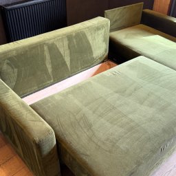 Górska Opieka Nieruchomości - Rozłożona sofa narożna z zieloną tapicerką welurową, ukazująca schowek. Widoczne ślady po praniu na siedzisku. Drewniana podłoga i dekoracyjna ściana w tle.
