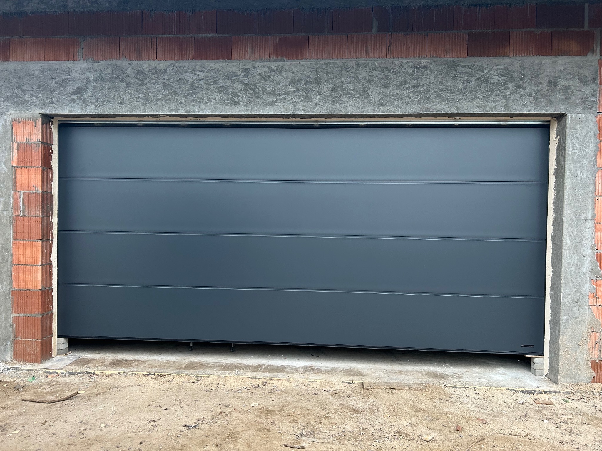 Szara segmentowa brama garażowa, widoczna od frontu w surowym otworze budynku z cegły i betonu. Brama zamknięta, z poziomymi przetłoczeniami.