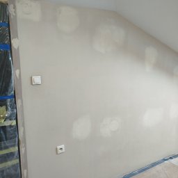 Ściana pod skosem po szpachlowaniu, widoczne gniazdko i włącznik. Zabezpieczona folią podłoga i framuga okienna. Etap przygotowania do malowania.