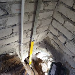 Kabel elektryczny w otworze w ścianie z cegły, obok rura PCV i metalowa listwa. Widoczny miernik. Praca w trakcie wykonywania, oświetlona punktowo.