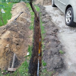 Wykopany wąski rów w ziemi, prawdopodobnie pod kable elektryczne, z widocznymi przewodami i narzędziami (młotek, łopata) obok zaparkowanego samochodu.