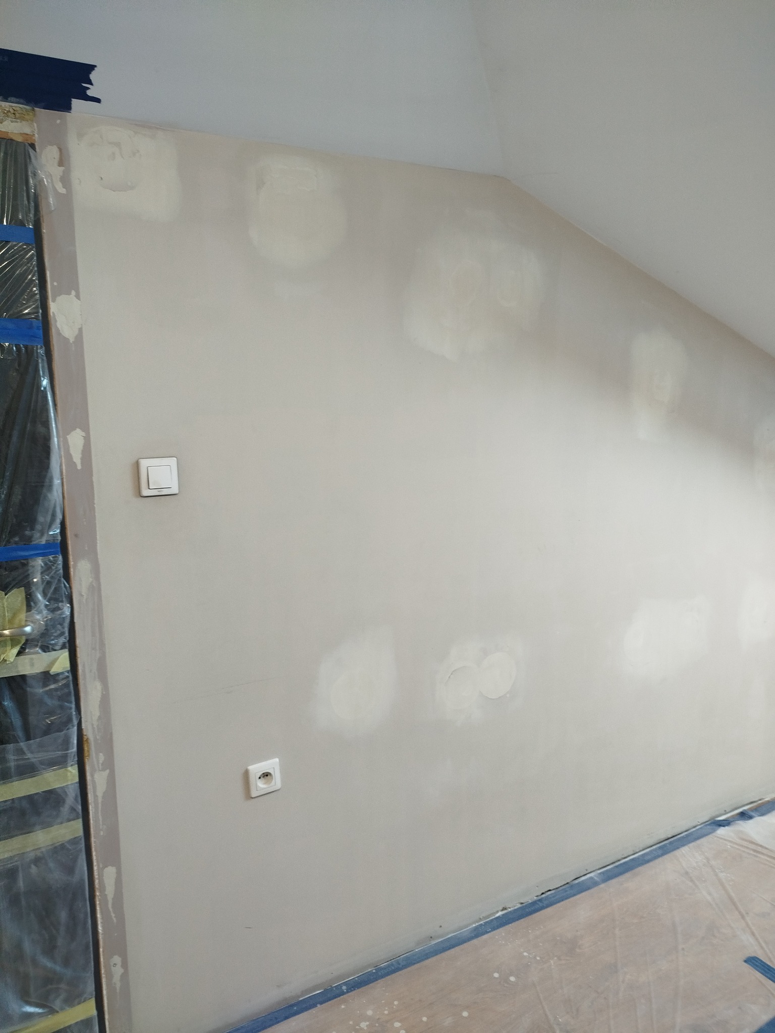 Ściana pod skosem po szpachlowaniu, widoczne gniazdko i włącznik. Zabezpieczona folią podłoga i framuga okienna. Etap przygotowania do malowania.