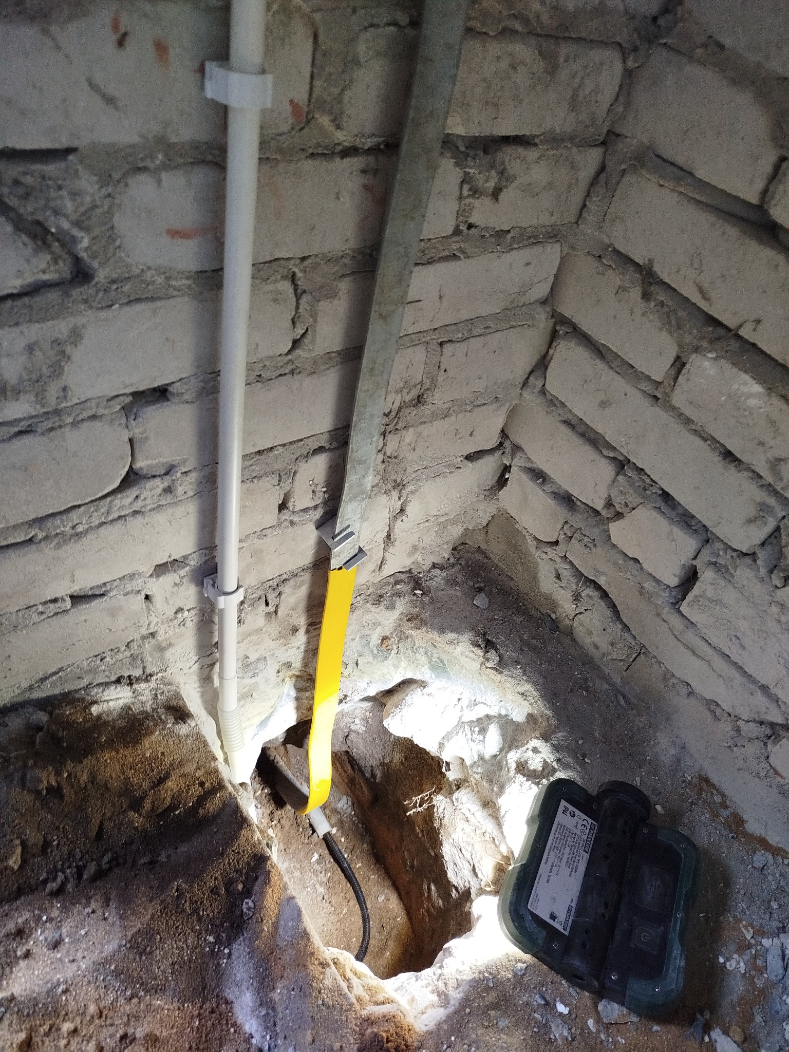 Kabel elektryczny w otworze w ścianie z cegły, obok rura PCV i metalowa listwa. Widoczny miernik. Praca w trakcie wykonywania, oświetlona punktowo.