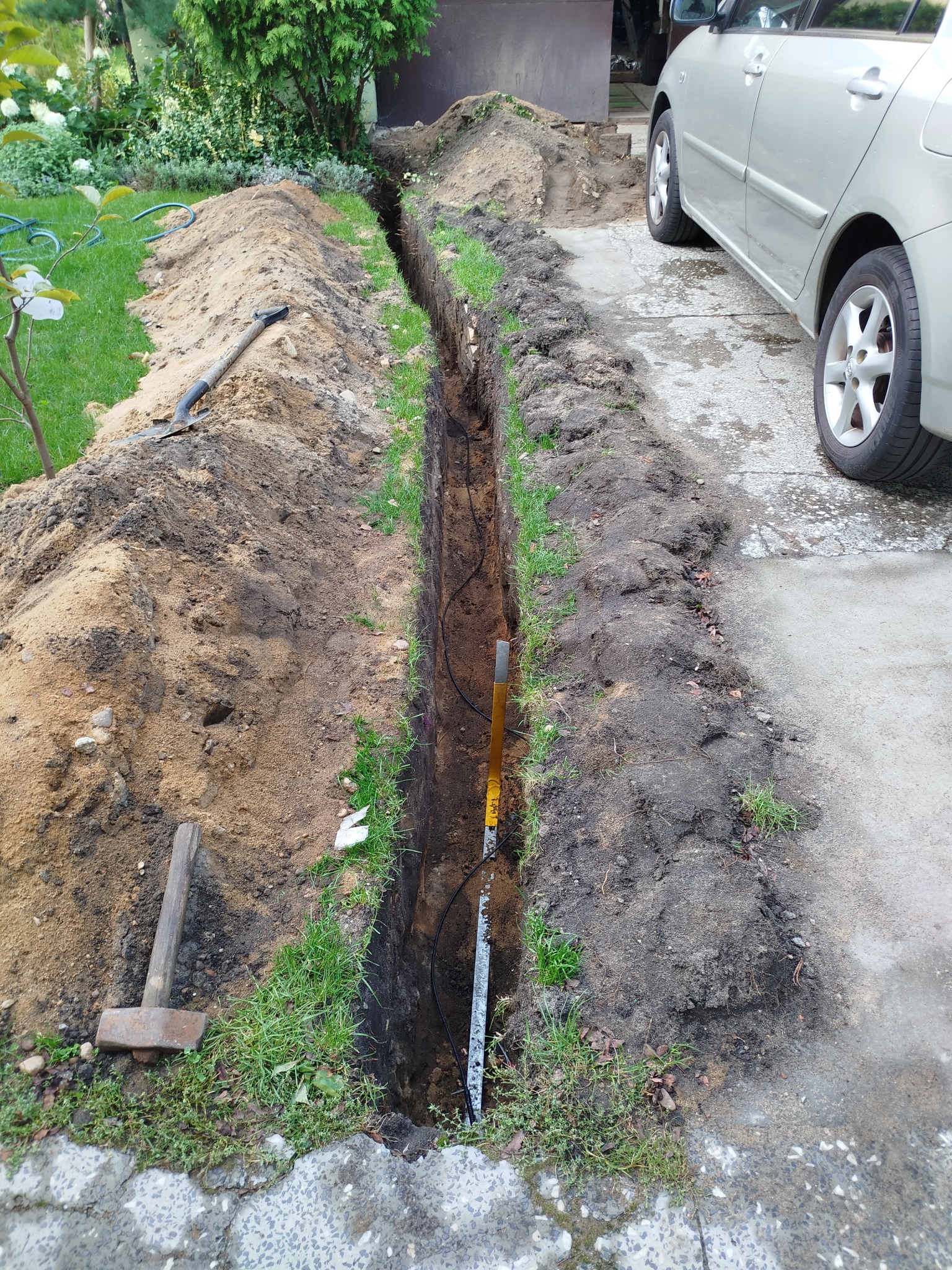 Wykopany wąski rów w ziemi, prawdopodobnie pod kable elektryczne, z widocznymi przewodami i narzędziami (młotek, łopata) obok zaparkowanego samochodu.
