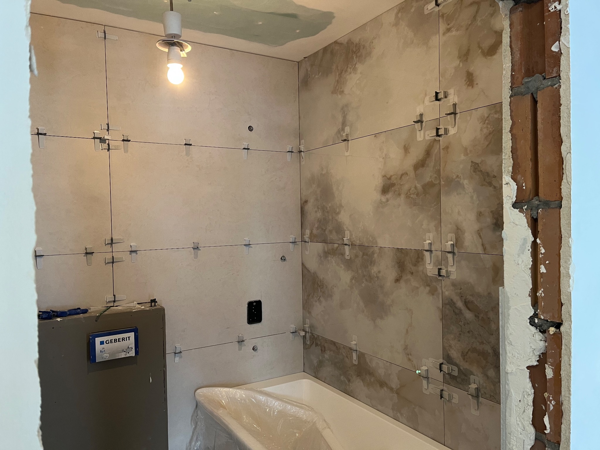 Układanie płytek w łazience z użyciem systemu poziomowania. Widoczna wanna zabezpieczona folią i fragment ściany z cegły. Oświetlenie sufitowe.