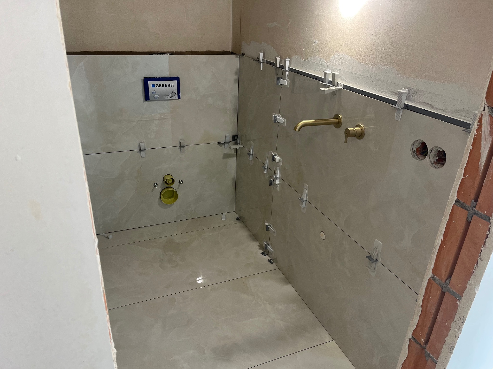 Beżowe płytki imitujące marmur, ułożone w łazience z widocznymi elementami instalacji i poziomowania. Złota bateria umywalkowa dodaje elegancji.