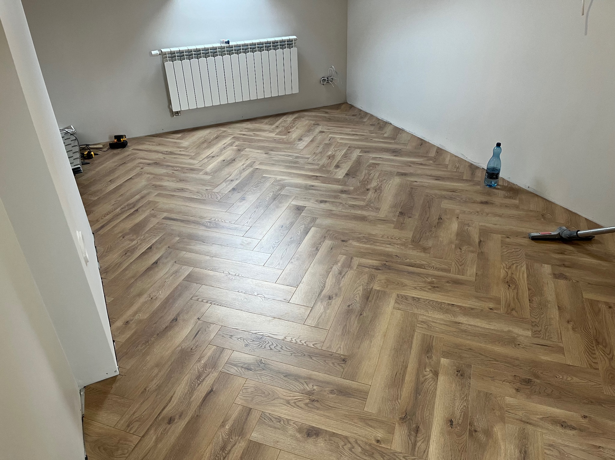 Ukończona podłoga z paneli ułożonych w jodełkę w jasnym pokoju z białymi ścianami i grzejnikiem. Widoczna butelka wody i odkurzacz.
