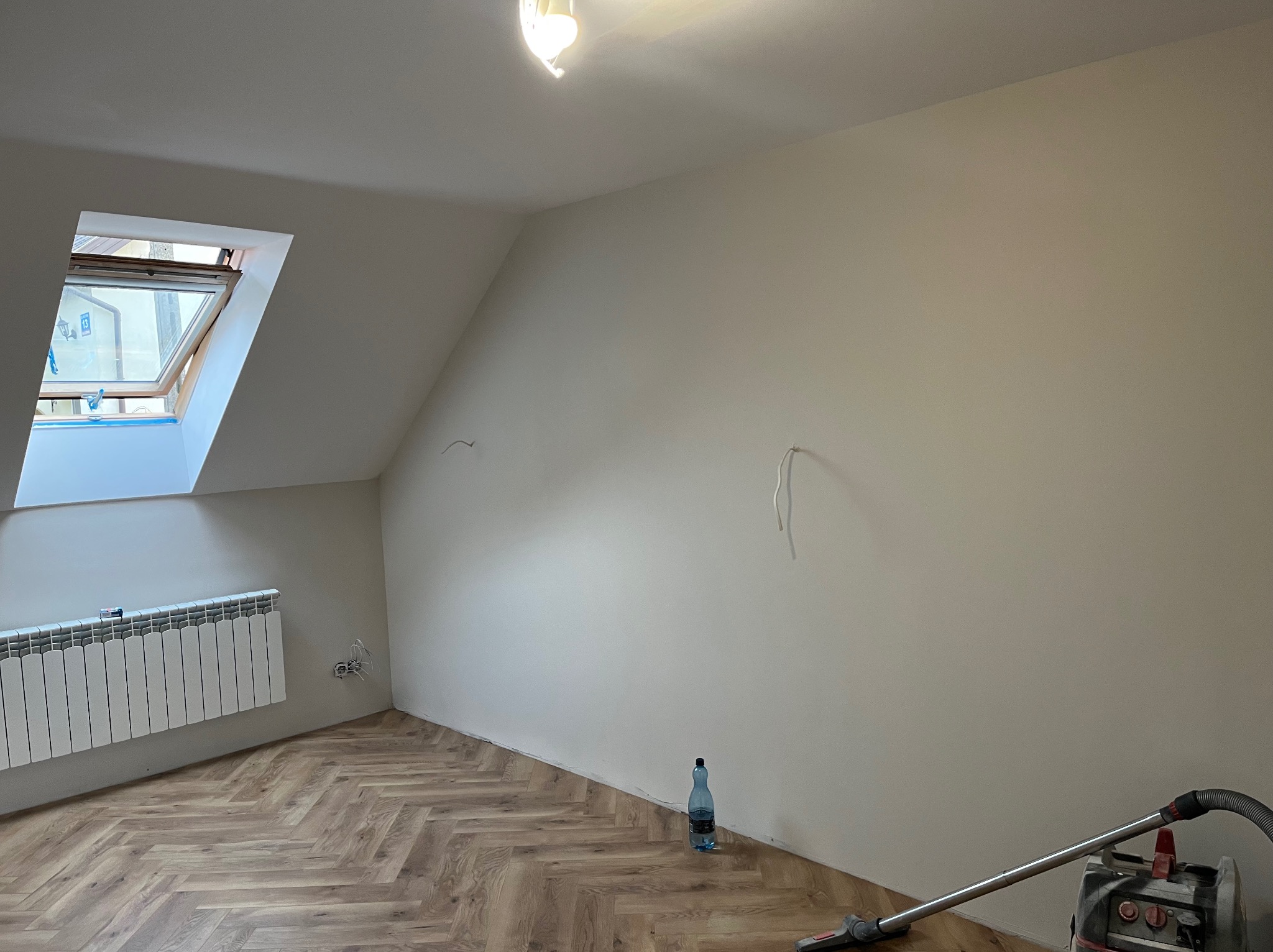 Poddasze po malowaniu ścian na jasny kolor, z nową podłogą w jodełkę. Widoczne okno dachowe, grzejnik, butelka wody i odkurzacz. Przygotowanie do dalszych prac wykończeniowych.