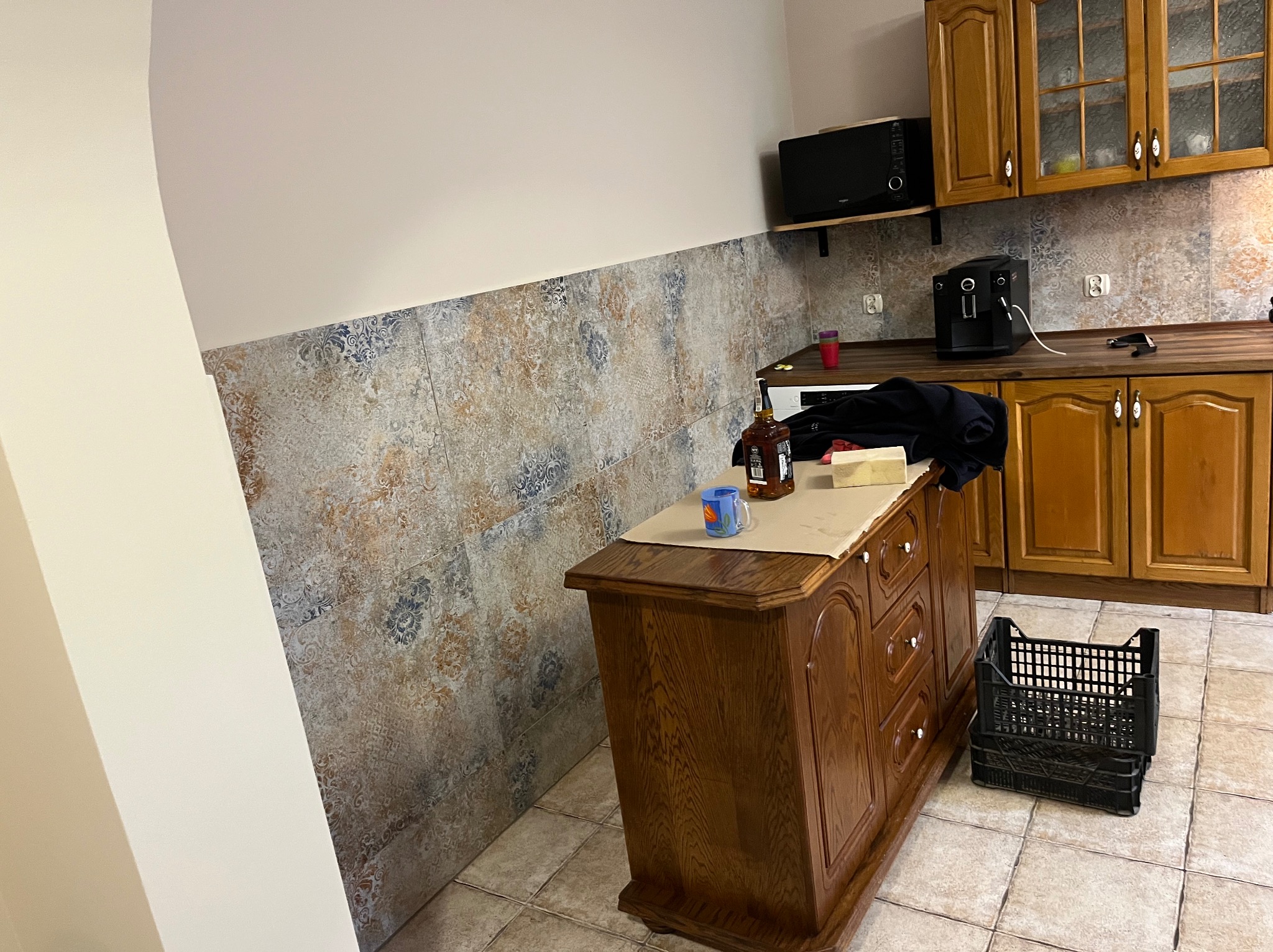 Kuchnia z drewnianymi szafkami, wzorzystą ścianą i wyspą. Na blacie wyspy kubek i butelka. Na podłodze kratka. Widok na fragment wykończonego wnętrza.