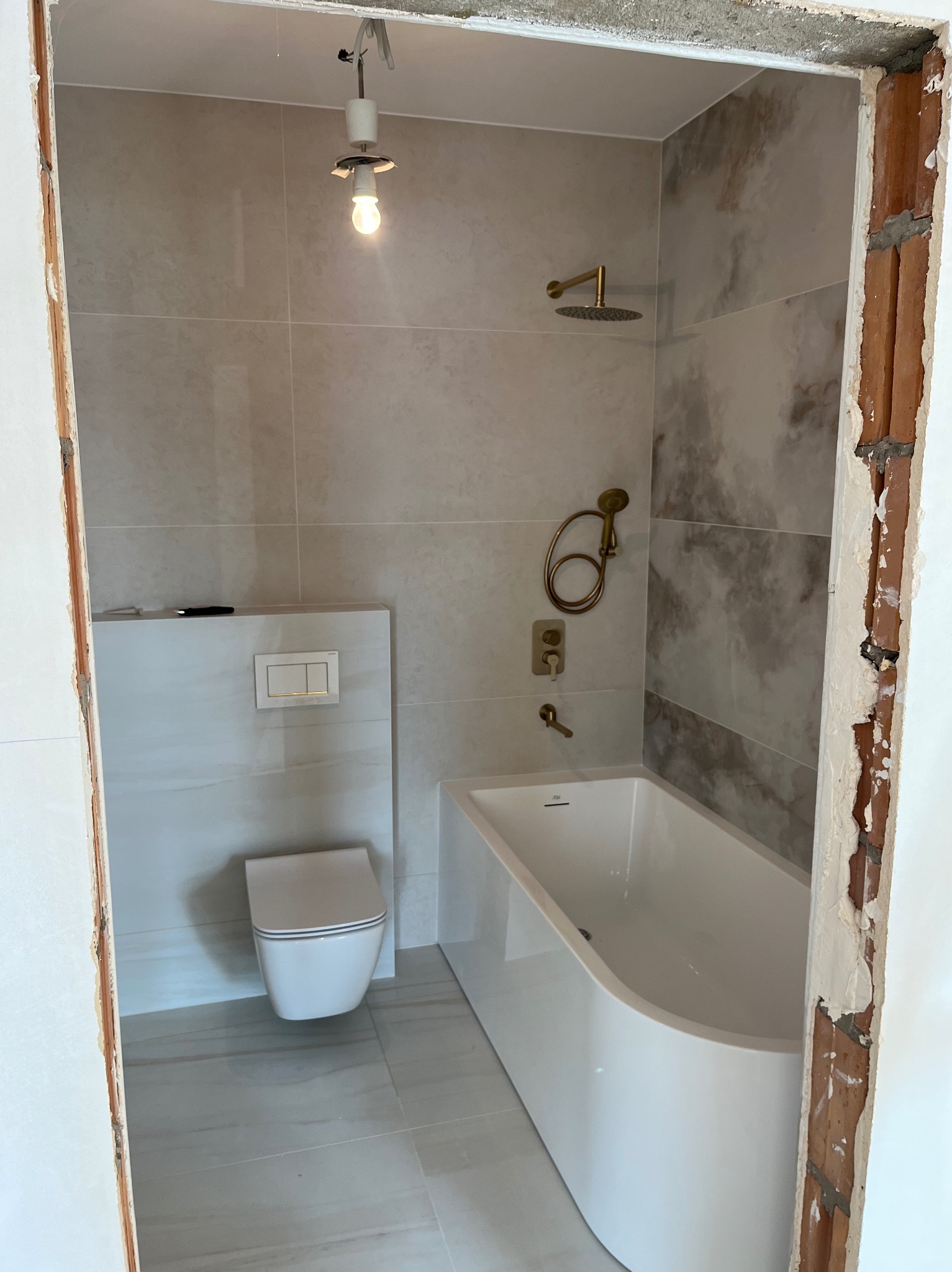 Wykończona łazienka z beżowymi płytkami, złotą armaturą i podwieszaną toaletą. Widoczny surowy fragment ściany przy wejściu, podkreślający etap prac wykończeniowych.