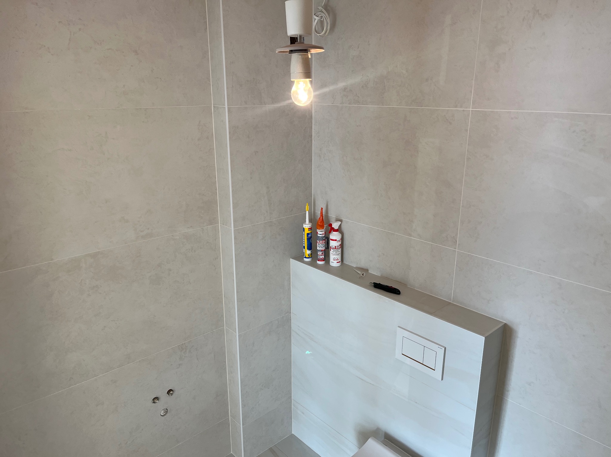 Wykończona łazienka z jasnymi, kwadratowymi płytkami. Na zabudowie stelaża WC stoją silikony i nożyk. Wisząca żarówka oświetla wnętrze. Widoczne otwory na baterię.