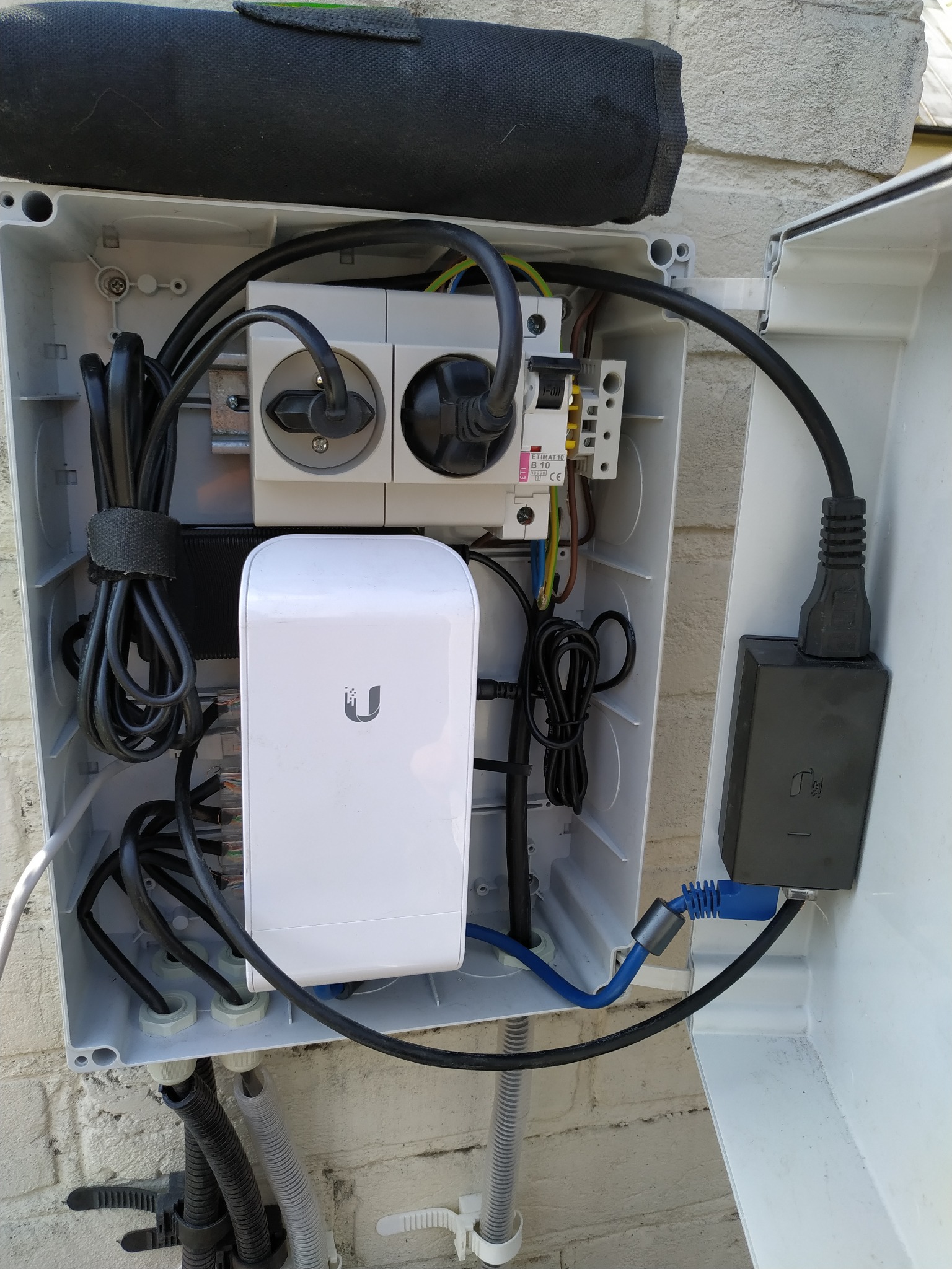 Otwarta skrzynka z elementami instalacji elektrycznej i routerem WiFi, widoczne kable, gniazdka i zabezpieczenia. Skrzynka zamontowana na ścianie budynku.