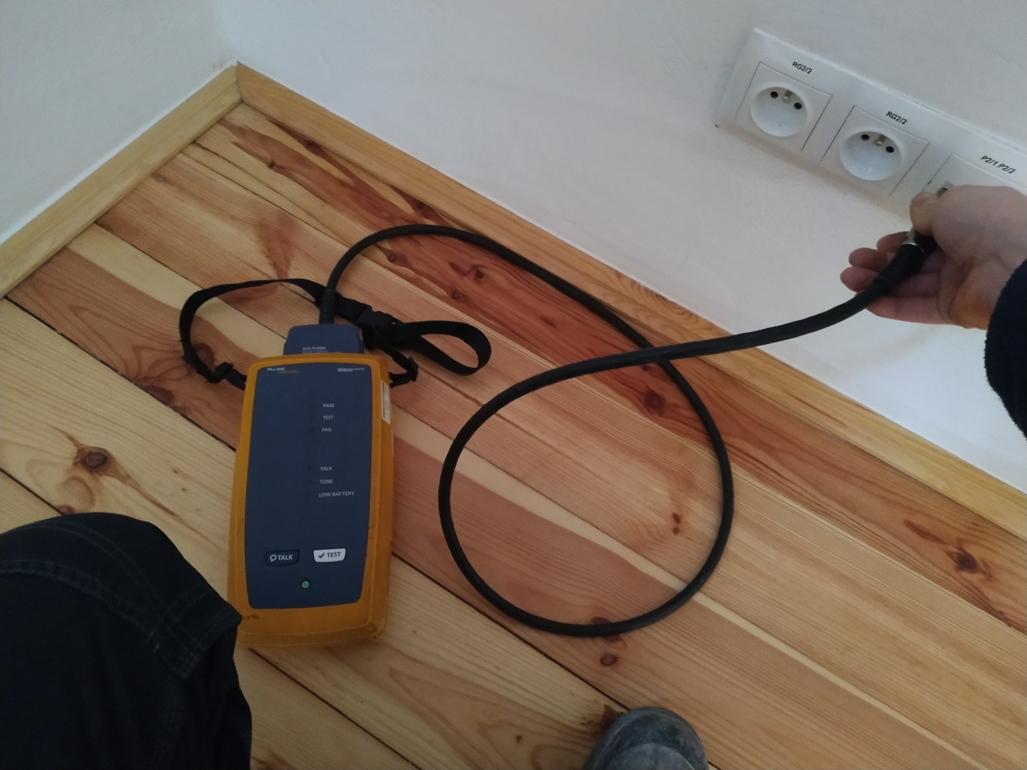 Testowanie gniazdka antenowego testerem kabli na drewnianej podłodze. Widoczny tester w żółtej obudowie, kabel i dłoń wkładająca wtyczkę do gniazdka.