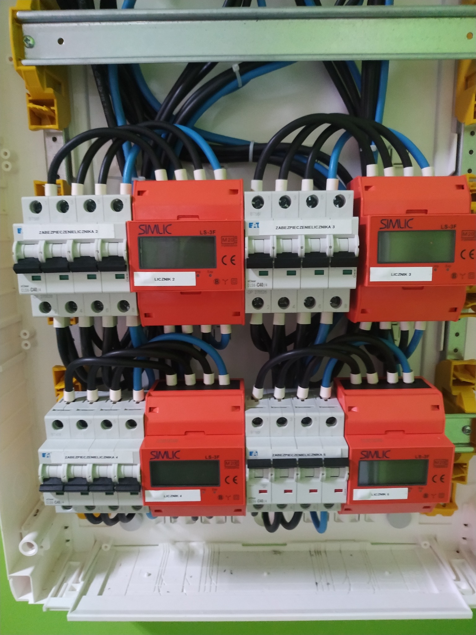 Rozdzielnica elektryczna z bezpiecznikami i przewodami w kolorach czarnym i niebieskim, zamontowana w białej obudowie. Widoczne elementy sterujące i oznaczenia.