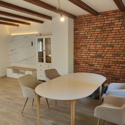 KEVINSON - Jasne wnętrze jadalni z ceglaną ścianą, drewnianymi belkami stropowymi i nowoczesnymi meblami. Widoczna szafka, stół i krzesła w neutralnych barwach.