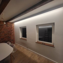 KEVINSON - Jasne wnętrze po remoncie: białe ściany z oknami, drewniane belki sufitowe z oświetleniem LED, ceglana ściana, drewniana podłoga i biały stół. Widok na odnowione pomieszczenie.