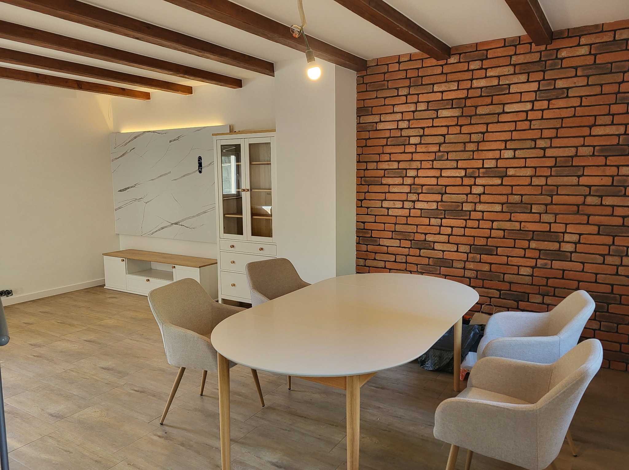 Jasne wnętrze jadalni z ceglaną ścianą, drewnianymi belkami stropowymi i nowoczesnymi meblami. Widoczna szafka, stół i krzesła w neutralnych barwach.