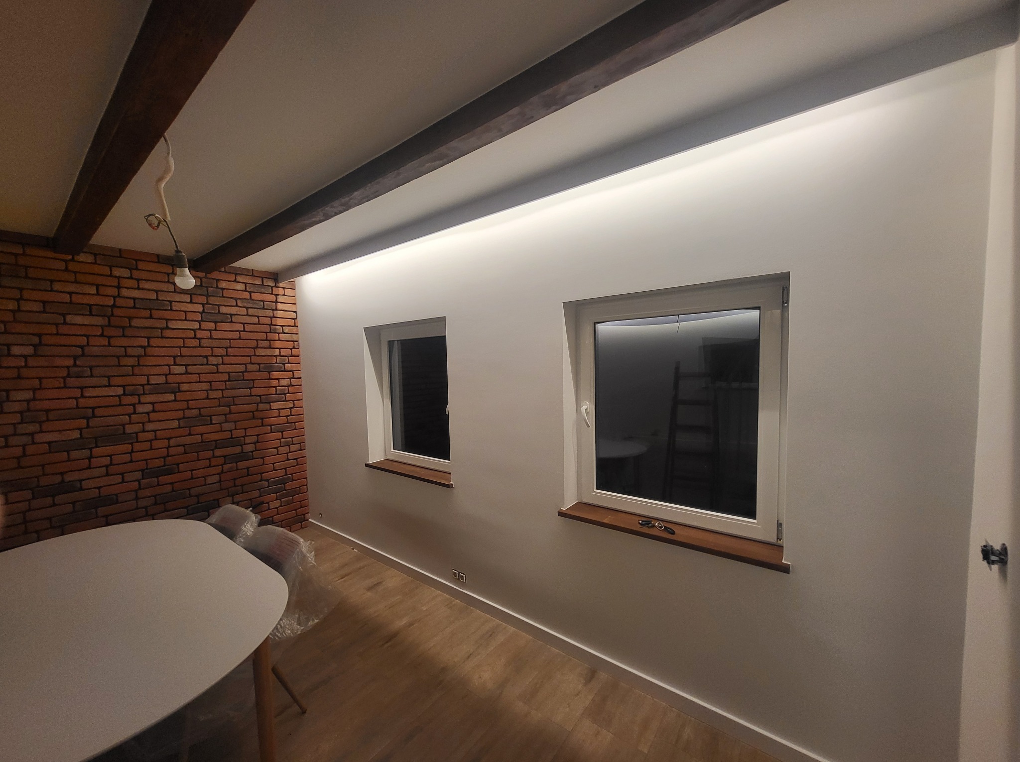 Jasne wnętrze po remoncie: białe ściany z oknami, drewniane belki sufitowe z oświetleniem LED, ceglana ściana, drewniana podłoga i biały stół. Widok na odnowione pomieszczenie.