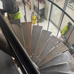 Kamil Rekowski - Spiralne schody z metalowymi stopniami w trakcie montażu. Widoczny pracownik z narzędziem, wnętrze domu z oknem i kominkiem w tle. Ujęcie z góry.