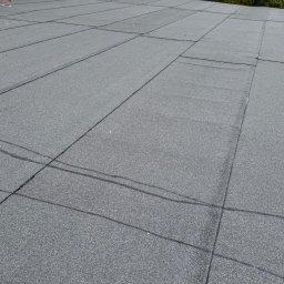 Szczegółowe ujęcie płaskiego dachu pokrytego szarą papą bitumiczną w kwadratowych panelach, z widocznymi liniami łączeń i fragmentem budynku w tle.