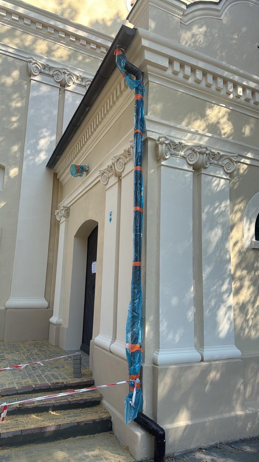 Elewacja budynku z ozdobnymi detalami architektonicznymi. Rynna zabezpieczona folią i taśmą malarską podczas prac renowacyjnych. Widoczne schody wejściowe i fragment okna.