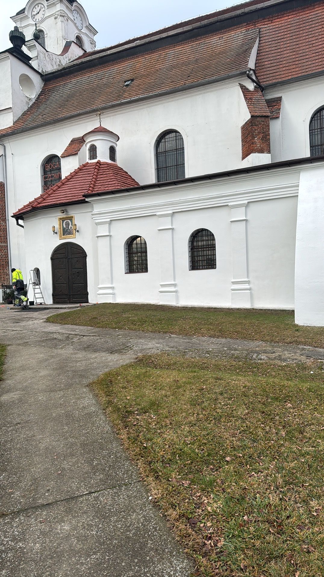 Malowanie elewacji kościoła na biało. Pracownik na drabinie, widoczny fragment budynku z czerwonym dachem i elementami architektonicznymi. Prace w toku.