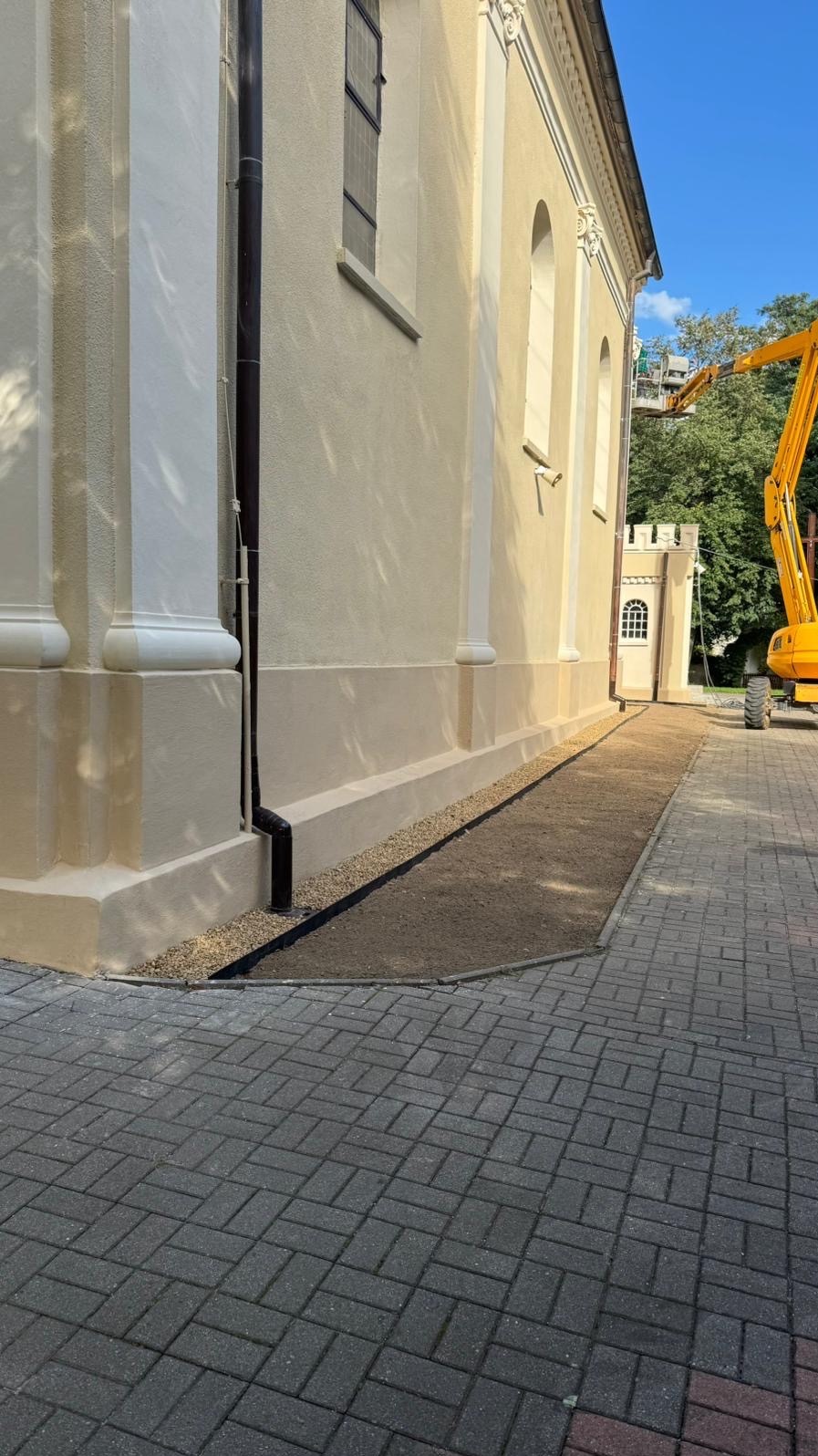 Odnowiona elewacja budynku z kolumnami i nowym systemem odprowadzania wody. Widoczny fragment chodnika z kostki brukowej i maszyna budowlana w tle.