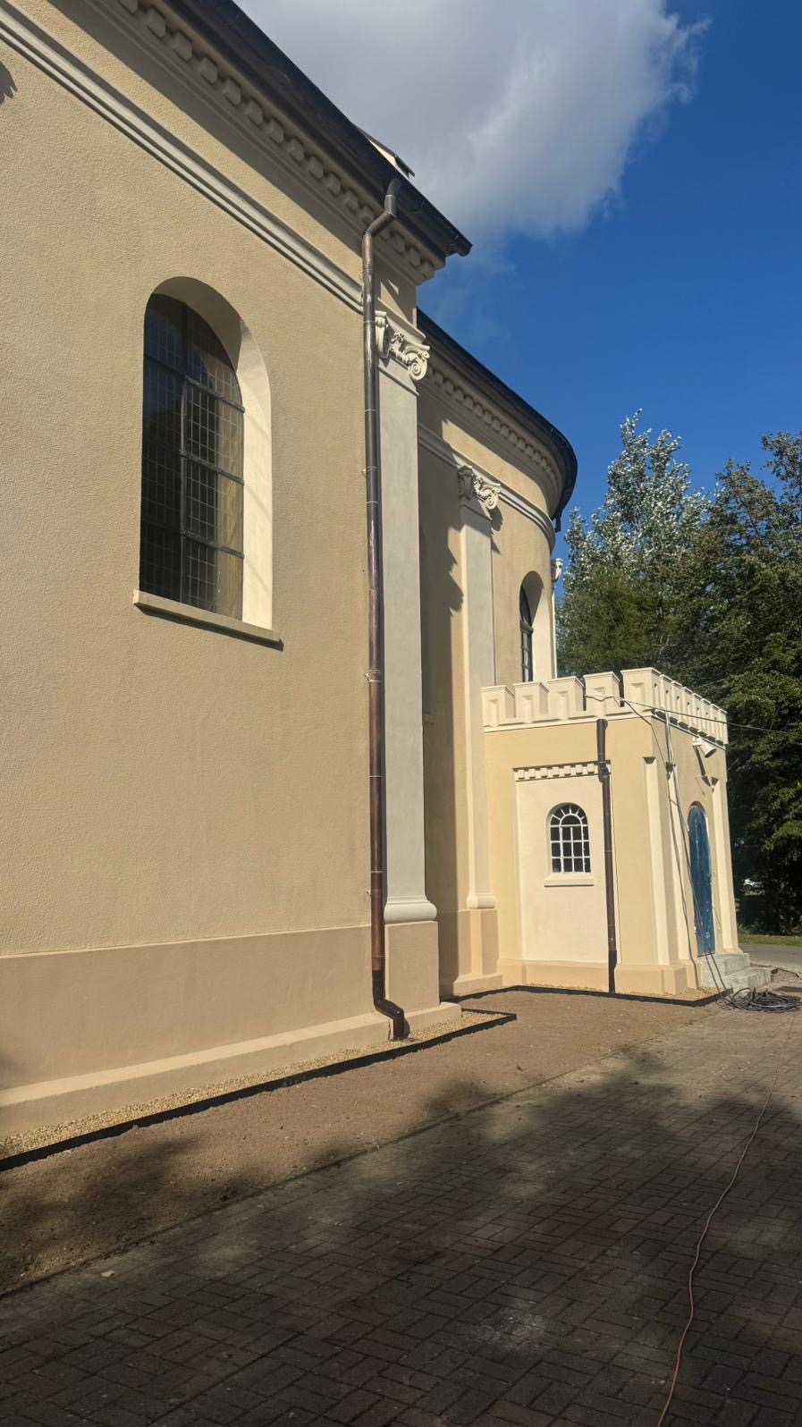 Odnowiona elewacja kościoła w ciepłych, beżowych tonacjach. Widoczne detale architektoniczne, okna z łukami i świeżo pomalowane ściany. Ujęcie z dołu, podkreślające wysokość budynku.