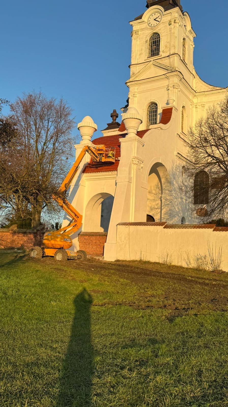 Prace na dachu kościoła z użyciem podnośnika koszowego. Czerwona dachówka, biała fasada, błękitne niebo. Widoczny cień osoby robiącej zdjęcie.