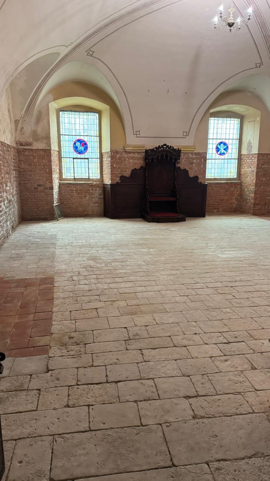 Sala z ceglaną ścianą, kamienną podłogą i ozdobnym krzesłem. Dwa okna z witrażami. Widoczne ślady renowacji podłogi. Wnętrze w trakcie prac wykończeniowych.