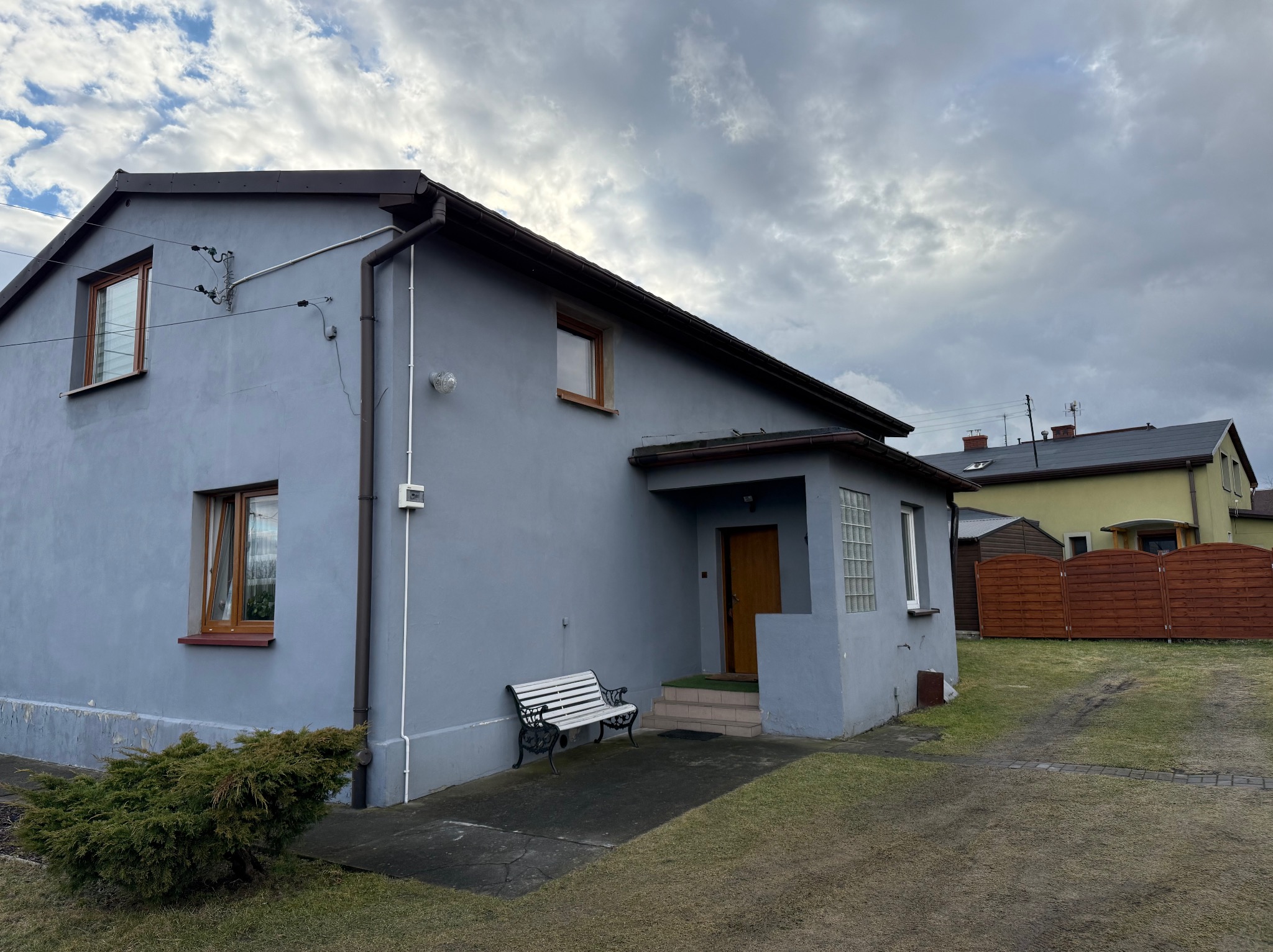 Niebieski dom jednorodzinny z brązowymi detalami, widoczny z boku, z ławeczką przed wejściem i pochmurnym niebem w tle. Widoczne instalacje na elewacji.