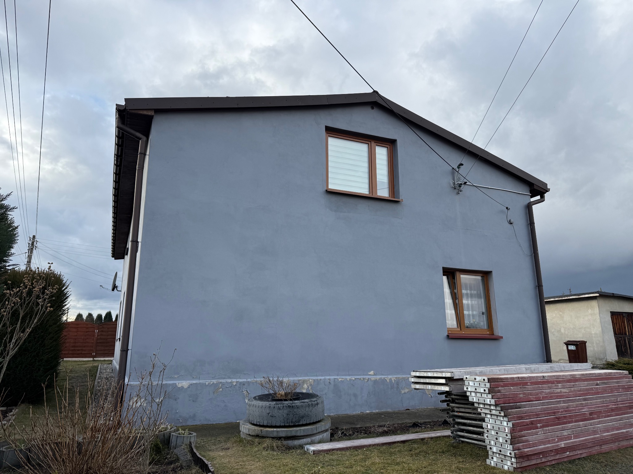 Szara elewacja domu jednorodzinnego z brązowymi oknami i dachem, widok z boku, z elementami rusztowania i dekoracją z opon na trawniku. Pochmurne niebo w tle.