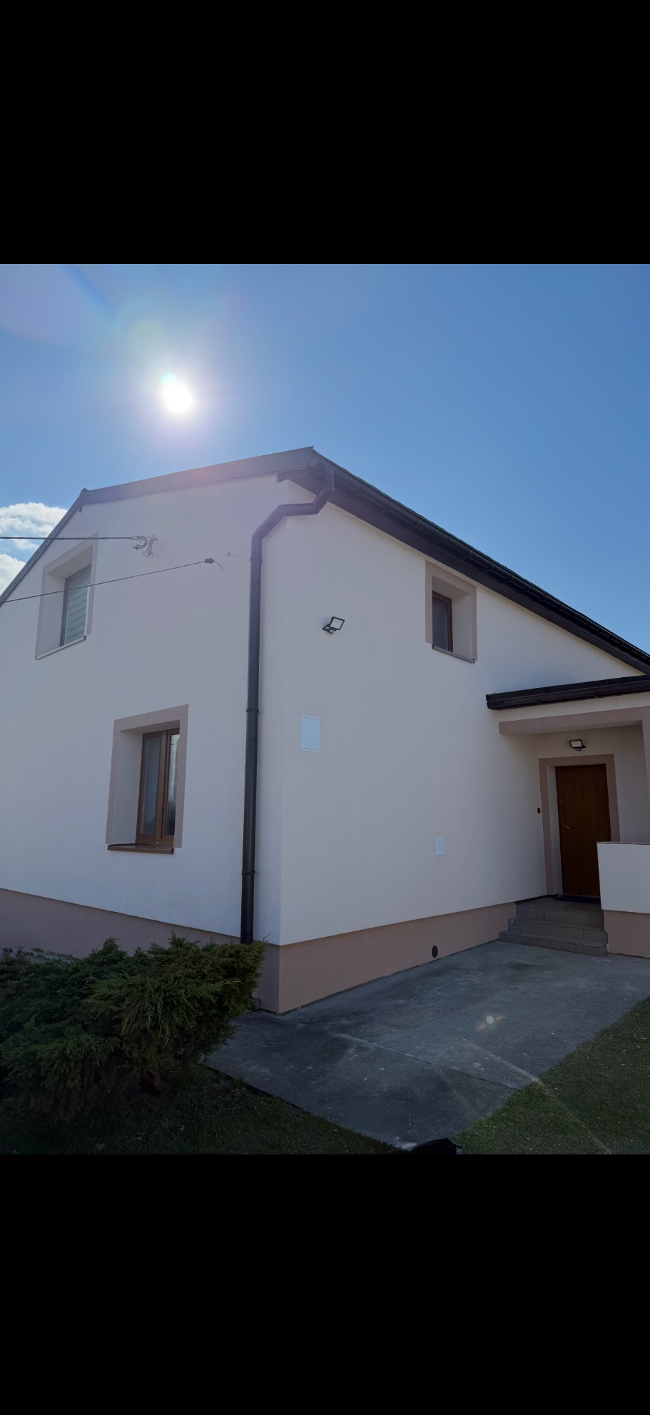 Jasny dom jednorodzinny po renowacji elewacji, z brązowymi oknami i rynnami, oświetlony słońcem na tle błękitnego nieba. Widoczny fragment ogrodu z krzewem.