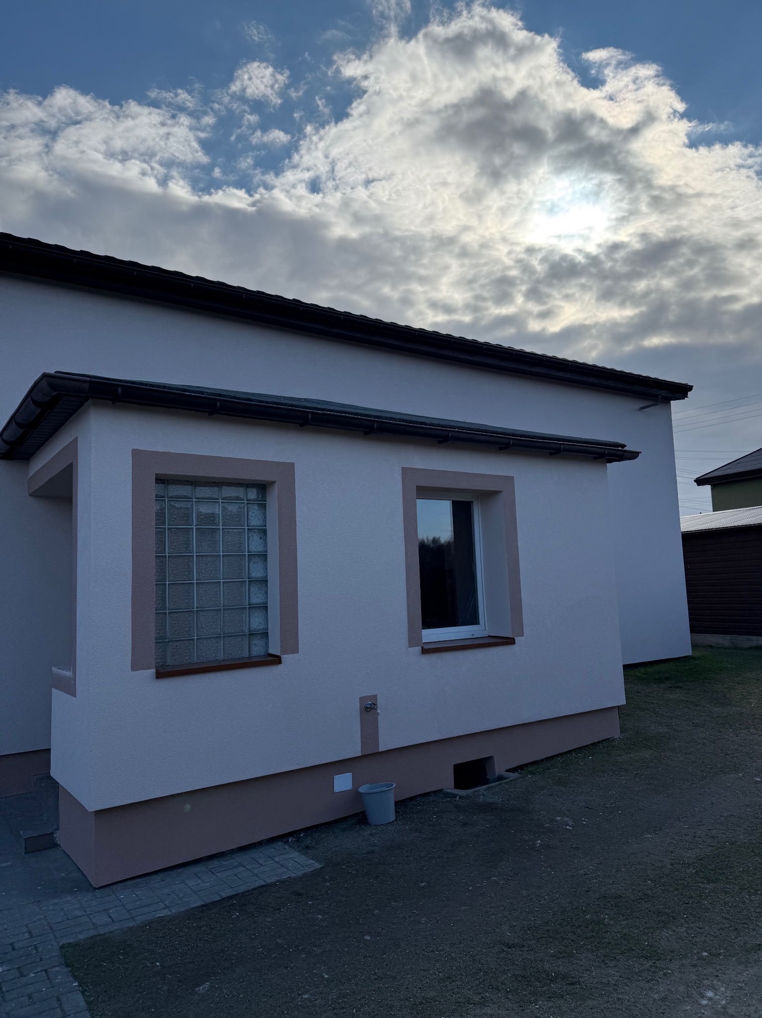 Otynkowany budynek z dwoma oknami, z których jedno jest z luksferów. Świeżo pomalowana elewacja w odcieniach bieli i beżu. Widoczne niebo z chmurami.